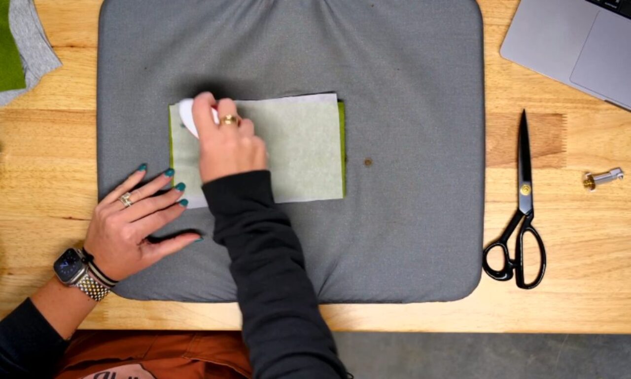 A person is using a small white iron to press a piece of fabric covered with parchment or transfer paper on a gray ironing pad. The person's left hand is holding the fabric in place. There are scissors, a sewing machine bobbin, and a laptop partially visible on the wooden table. The person is wearing a watch and several rings.