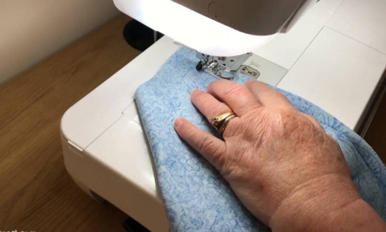 A person's hand guiding light blue fabric with a subtle floral pattern through a white sewing machine. The person is wearing two gold rings on their ring finger. The sewing machine is positioned on a wooden surface.