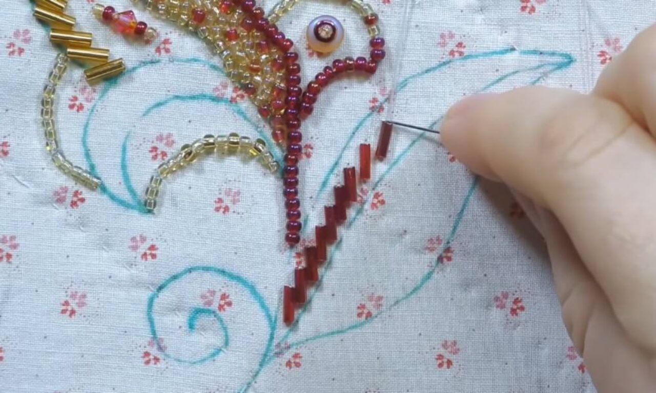 A close-up of a hand sewing red cylindrical beads onto fabric with a needle. The fabric has a floral print and a blue pattern drawn on it, with various beads in red, gold, and yellow forming a decorative design.