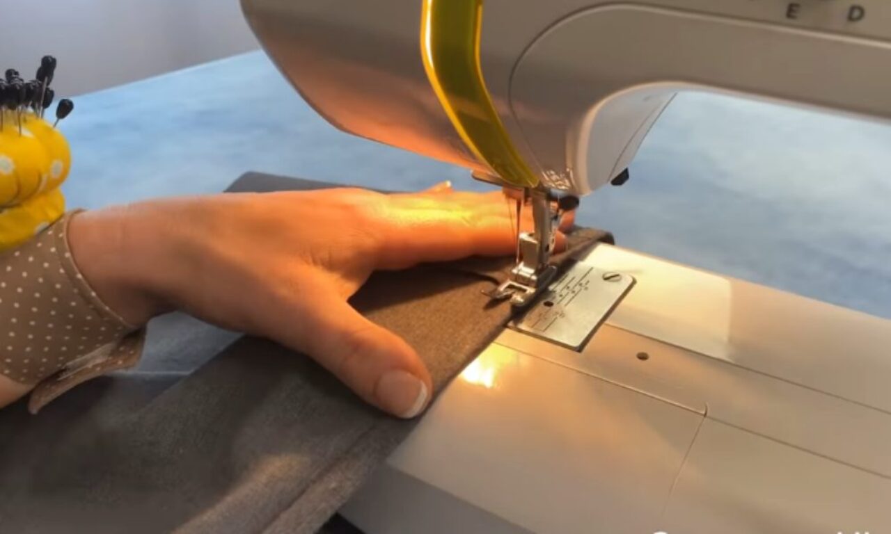 A close-up view of a person’s hands guiding grey fabric under the presser foot of a white sewing machine. The left wrist is fitted with a yellow pincushion, and a warm light illuminates the stitching area.