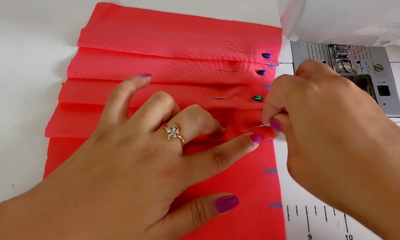 A close-up of hands folding and pinning pleats into a piece of bright pink fabric, preparing it for sewing on a sewing machine. The person's nails are painted purple, and they are wearing a flower-shaped ring on one finger. Multiple colorful pins hold the pleats in place along the edge of the fabric.