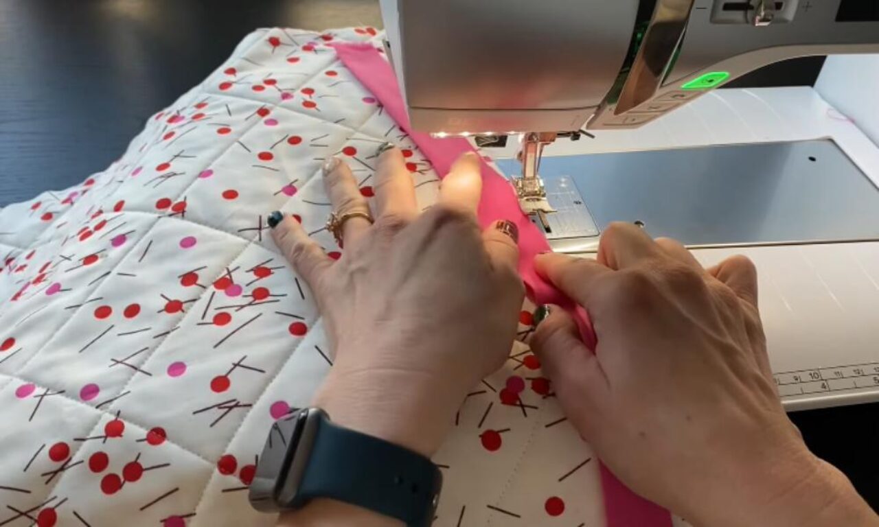 Hands sewing a pink fabric binding onto a quilted white fabric with a pattern of red and pink dots and black lines, using a sewing machine.