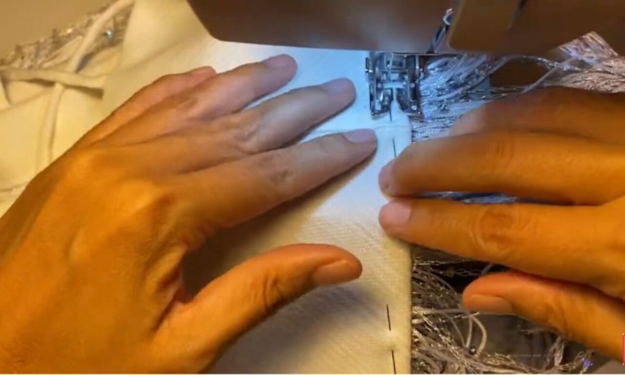 Close-up of two hands guiding a piece of white fabric through a sewing machine, with visible stitches along the edge.