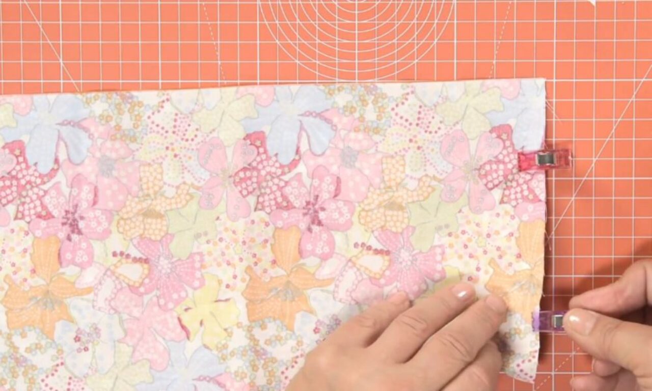 Hands pinning a piece of pastel, floral-patterned fabric with small clips on the right edge, laid on an orange gridded cutting mat.