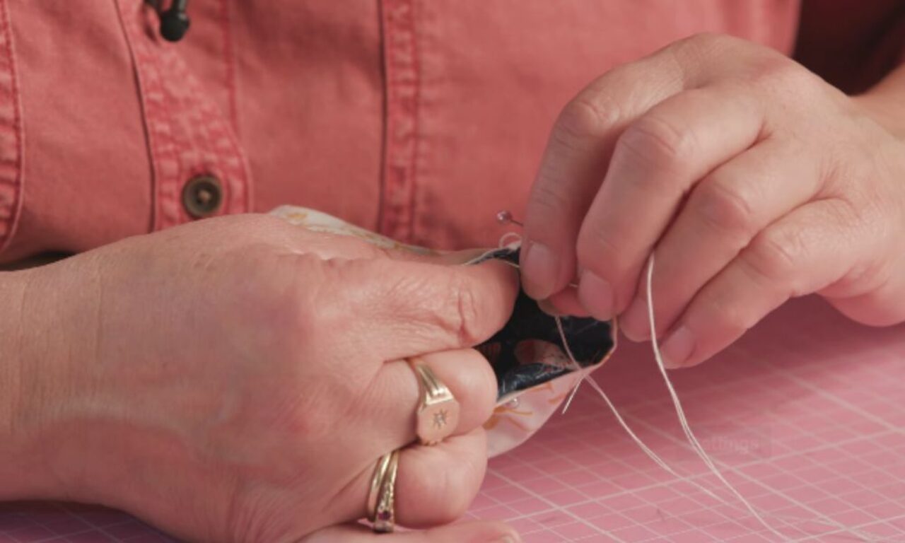 Close-up of a person hand-sewing two small pieces of patterned fabric; their fingers guide a needle threaded with white thread while they wear a salmon-colored button-down shirt and gold rings.