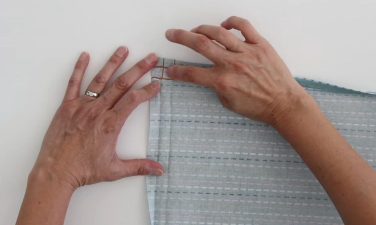 Hands folding the corner of light blue fabric with a dashed pattern, marking the fold.