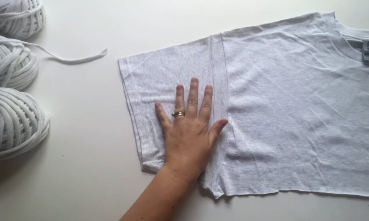 A hand placed on a white fabric, possibly a t-shirt, laid flat on a white surface. There are balls of thick white yarn visible on the left side.