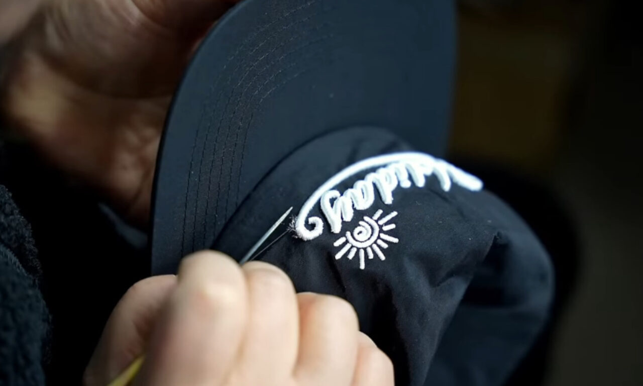 Hands using tweezers to trim excess threads from white embroidery on a black hat, showing the word "Holiday" and a sun symbol.
