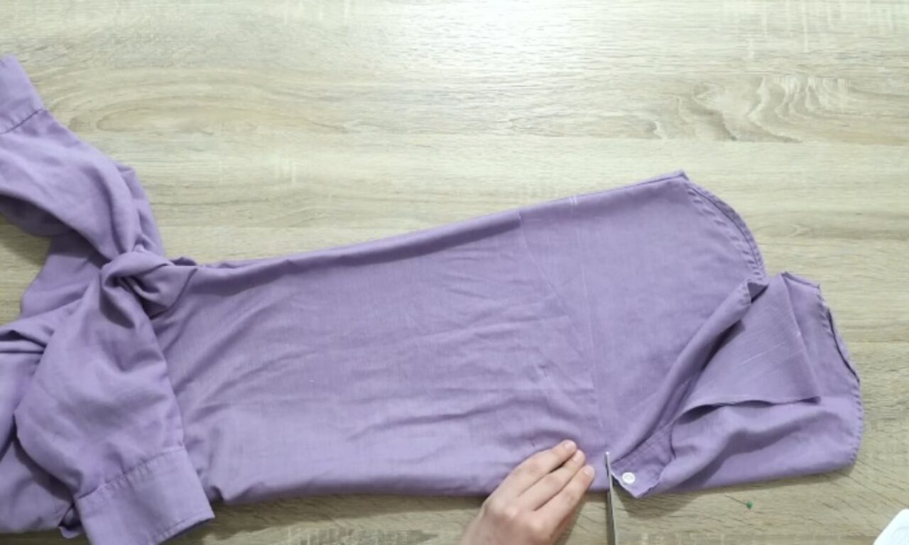 A person is cutting a lavender-colored fabric with scissors on a light wooden surface. The fabric appears to be part of a garment, possibly a pair of pants or a dress.