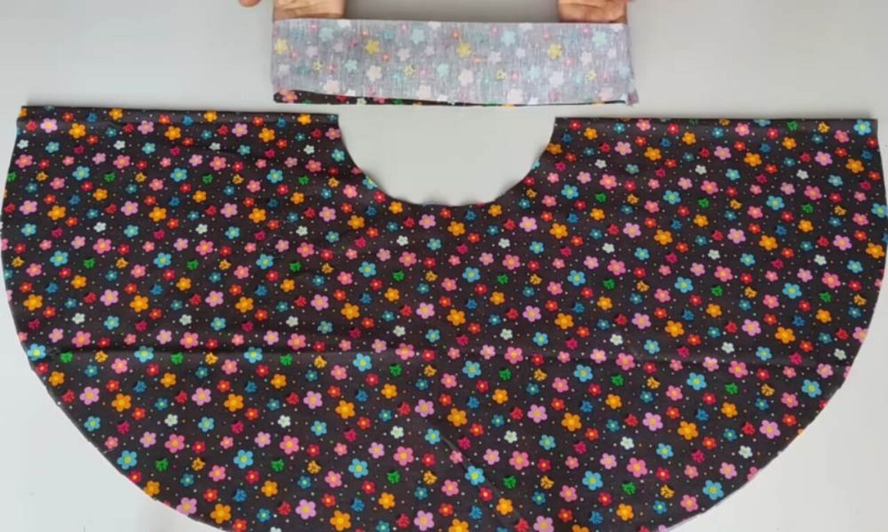 A large semicircular piece of black cotton fabric printed with small pink, orange, blue, and yellow flowers laid flat on a white surface, its straight edge featuring a U-shaped notch at center, and above it a narrow rectangular strip of light gray fabric with pastel star shapes held at its top corners by two hands.