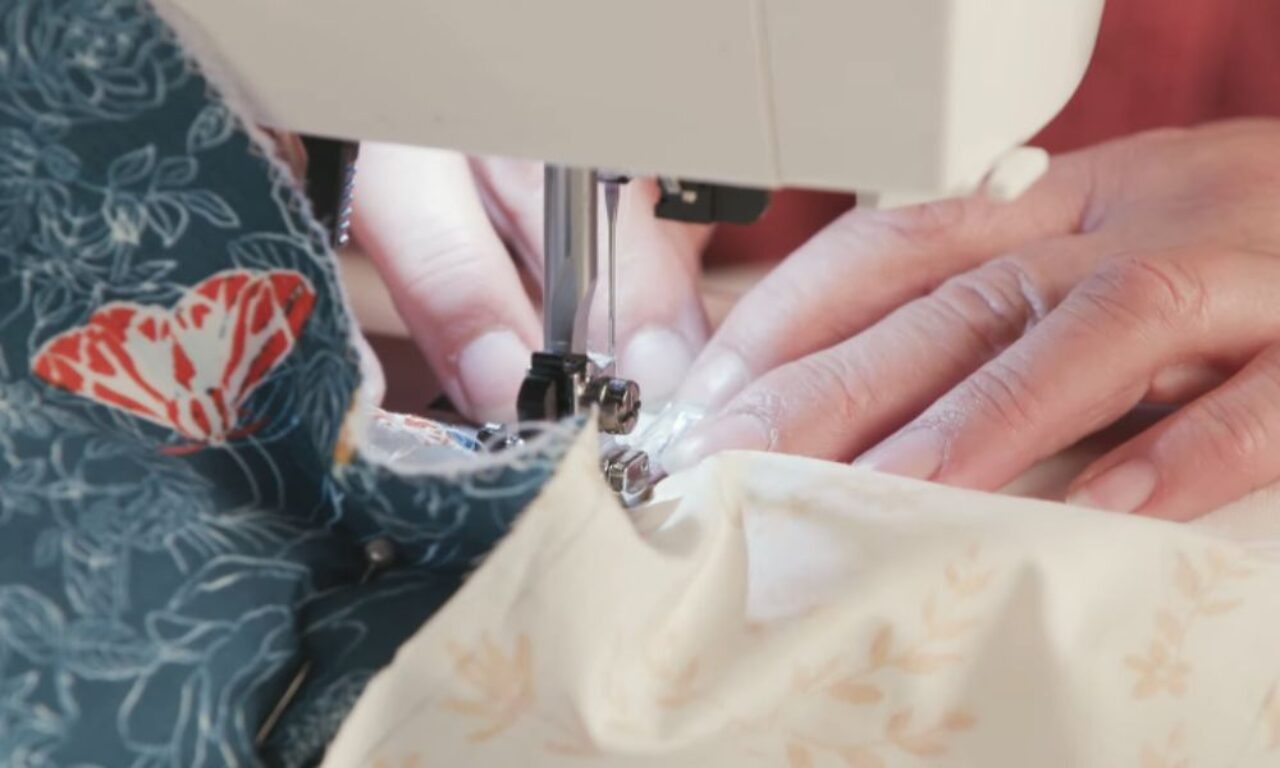 A close-up shot shows hands guiding two pieces of fabric under the needle of a sewing machine. One fabric is dark teal with a white floral outline and a prominent red and white butterfly design. The other is a light cream fabric with a subtle, lighter tan leaf pattern, being stitched along its edge.