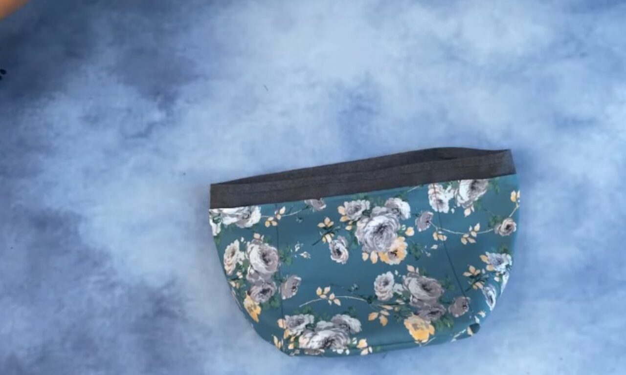 A floral fabric face mask with a dark gray trim at the top, featuring a pattern of white, gray, and yellow flowers on a teal background, placed on a light blue surface.