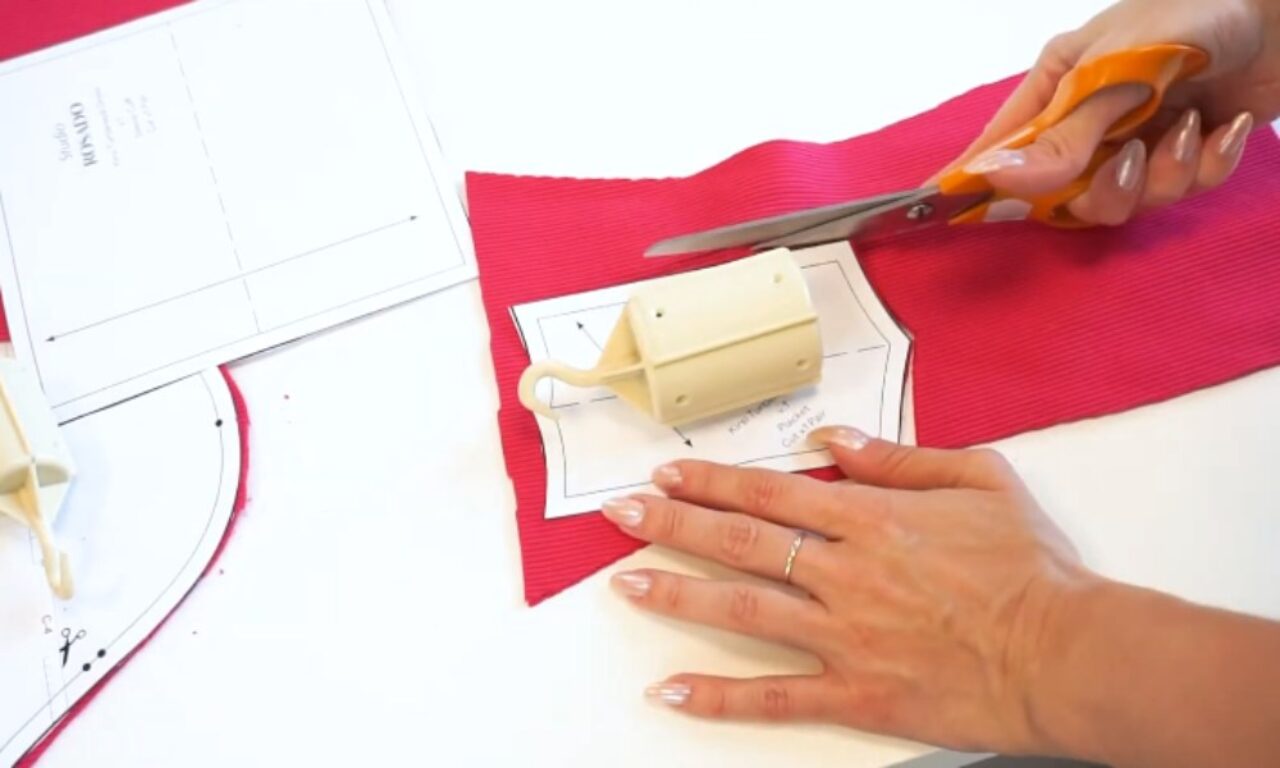 A person is cutting a piece of red fabric with orange-handled scissors. The fabric is placed on top of a pattern or template, which includes printed instructions and measurements. A beige rotary cutter is also visible near the fabric, suggesting that this is part of a sewing or crafting project. The person's hands are holding the scissors and stabilizing the fabric, indicating precision in the cutting process.
