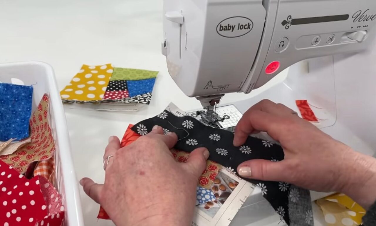 A close-up view of a pair of hands guiding a strip of black floral-print fabric through the needle of a white “Baby Lock” sewing machine. On the left, a plastic bin holds assorted colorful fabric scraps, and in the background several half-stitched▌