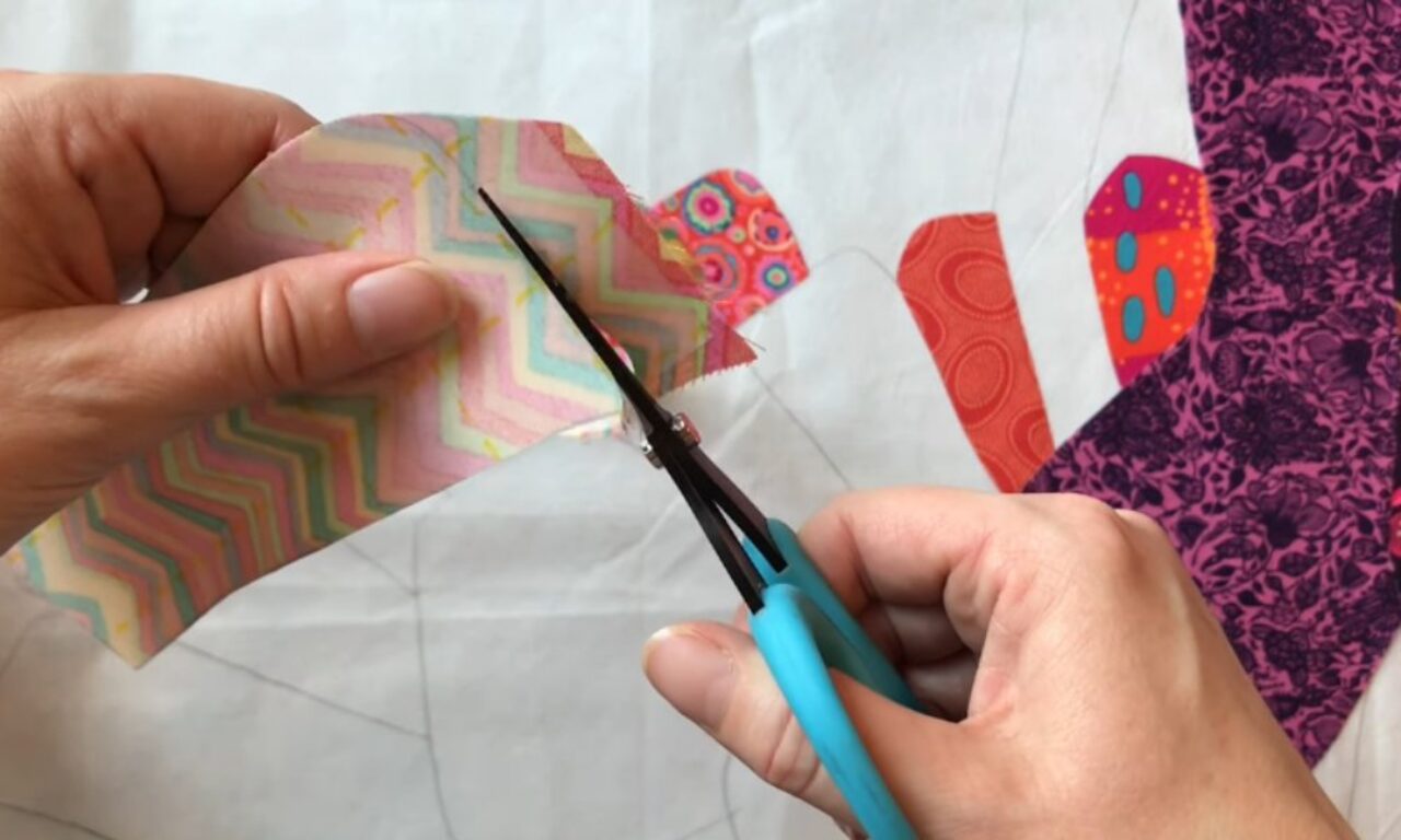 Two hands cutting a pastel chevron-patterned fabric shape with small blue-handled applique scissors, trimming along a faint pencil outline on a white background fabric sprinkled with other brightly colored appliqué pieces.