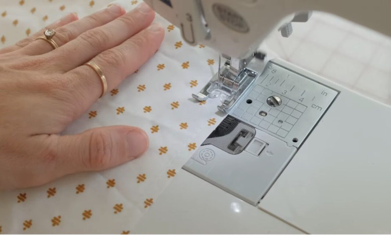A close-up of a sewing machine stitching white fabric with a small golden floral pattern. A person's left hand, adorned with two gold rings, guides the fabric under the presser foot. The metal plate of the sewing machine is marked with measurement lines in inches and centimeters.