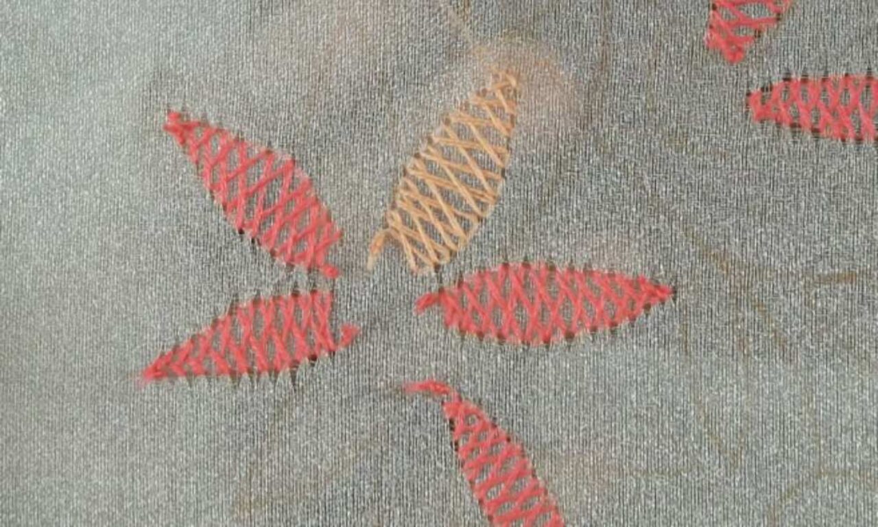 A close-up of textile embroidery featuring leaf-like shapes with cross-stitched patterns. Several coral-red leaf motifs and one tan/beige leaf are arranged on a gray fabric background. The embroidery has a diamond or lattice pattern within each leaf shape, creating a simple, decorative design with a handcrafted aesthetic.