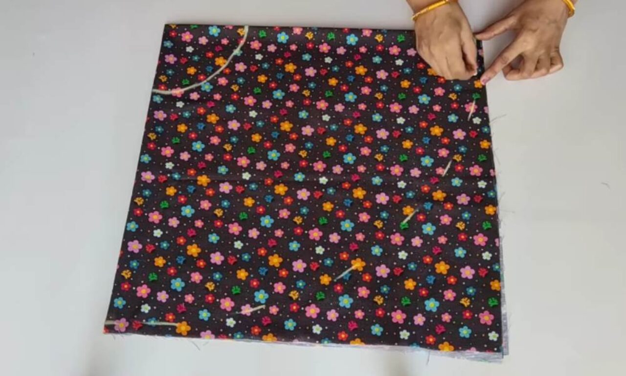 A person is holding a piece of black fabric with a colorful floral pattern against a white background. The fabric is adorned with small, multicolored flowers and is being grasped by two hands wearing gold bracelets, with loose threads visible along the edges.