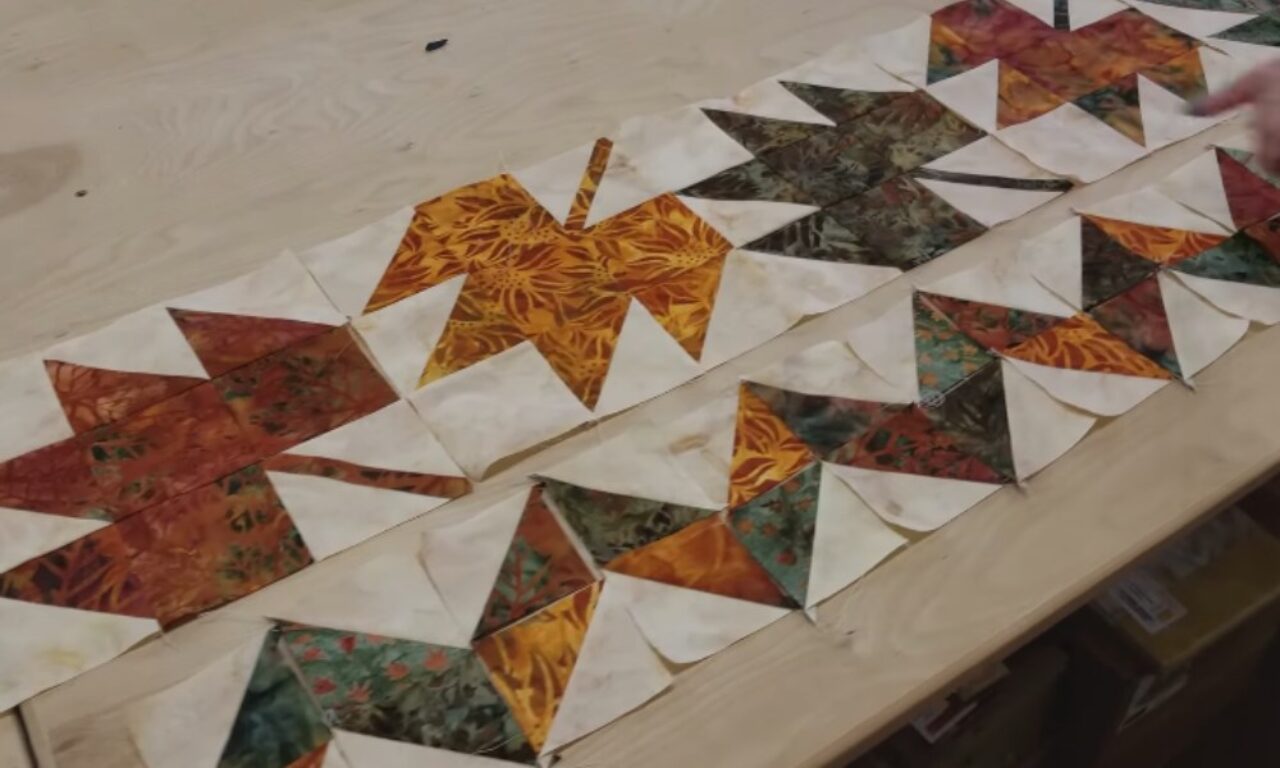 a quilt layout in progress, featuring a series of colorful, geometric blocks arranged on a light-colored wooden table.