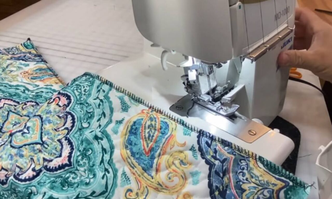 A close-up view of a sewing machine in action, with a hand guiding vibrant, paisley-patterned fabric as it stitches a colorful, zigzag trim along the edge. The machine sits on a white surface, surrounded by scattered threads and sewing tools, capturing a moment of creative productivity.