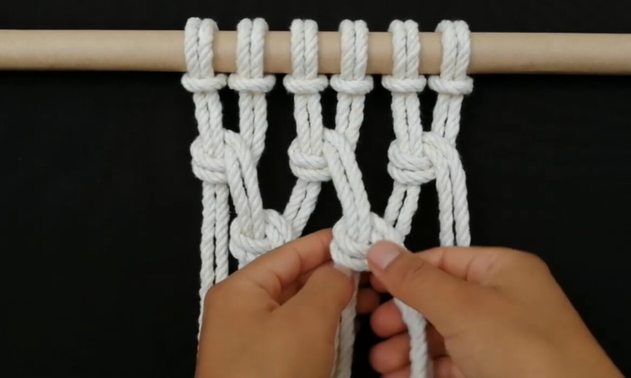 Hands are shown tying a knot in a white macrame rope project suspended from a wooden dowel against a black background. Several knots have already been formed in the hanging ropes.