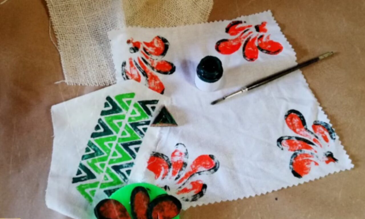 A white cloth with orange-and-black floral stamps and a green triangular pattern lies on a table, alongside a small jar of black paint, a paintbrush, a triangular print block, and a piece of burlap fabric.