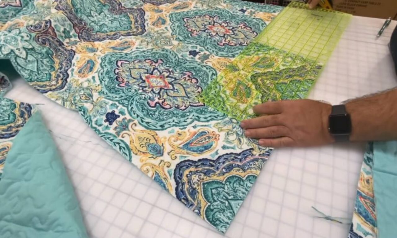 A person's hand, wearing a smartwatch, presses a transparent yellow gridded quilting ruler onto a colorful paisley and floral patterned fabric. The fabric, featuring shades of teal, blue, and yellow, is laid out on a white gridded cutting mat, with a piece of solid teal quilted material visible to the left, suggesting a sewing or quilting project.