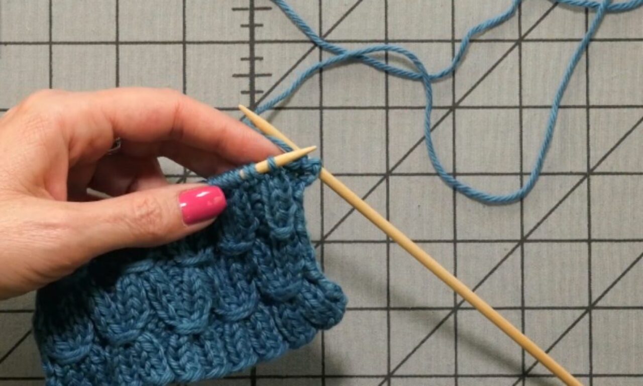 A close-up of a person’s left hand, with bright pink nail polish, holding a wooden knitting needle and a piece of blue knit fabric. The right-hand needle is inserted into a stitch, and a length of matching blue yarn loops across a gray cutting mat marked with a black grid.