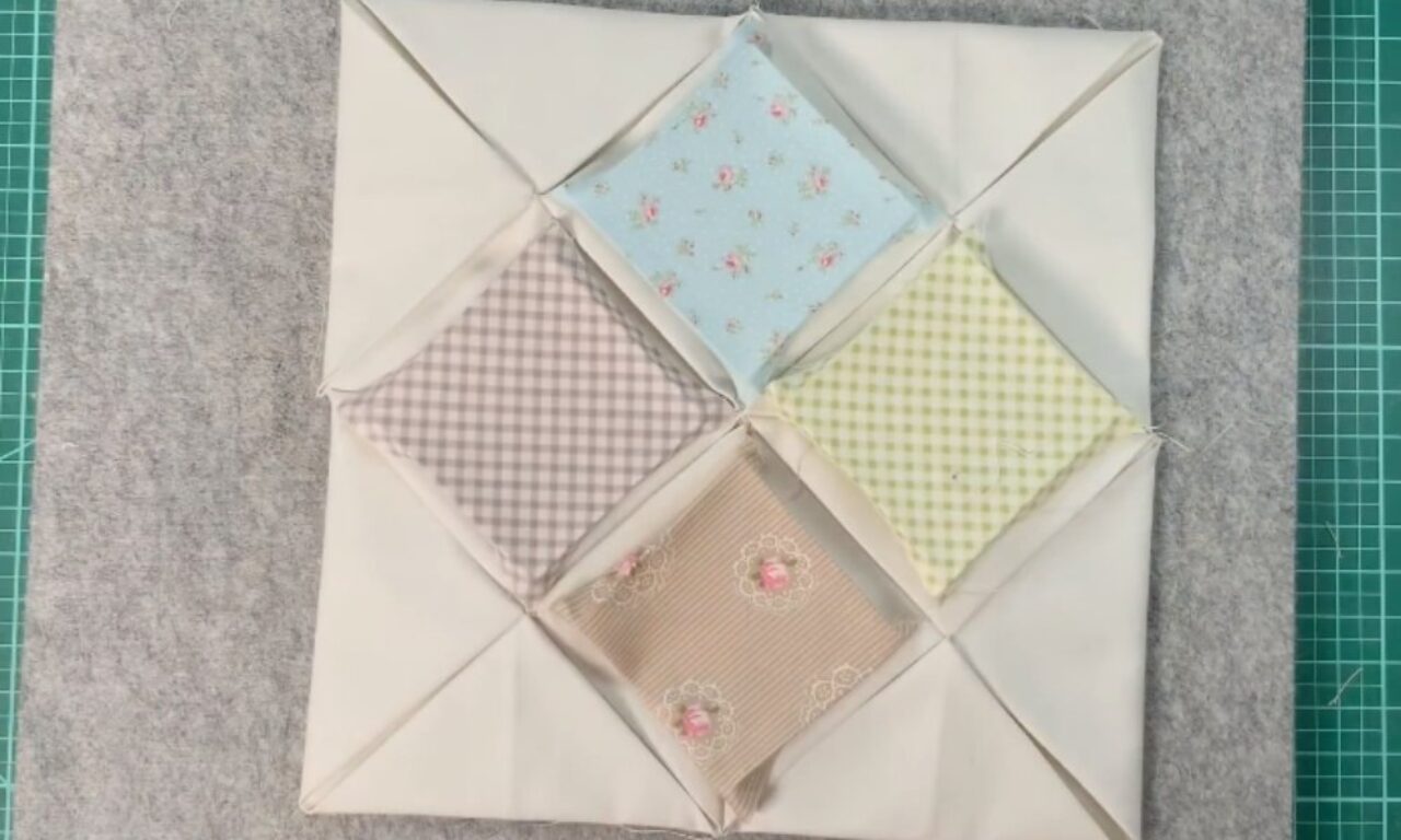 A square quilt block featuring four padded fabric squares arranged in a diamond shape. Each square has a pastel print—blue with tiny roses, gray gingham, green gingham, and beige with rose motifs—framed by white triangular borders. The block rests on a gray felt mat over a green grid cutting mat.