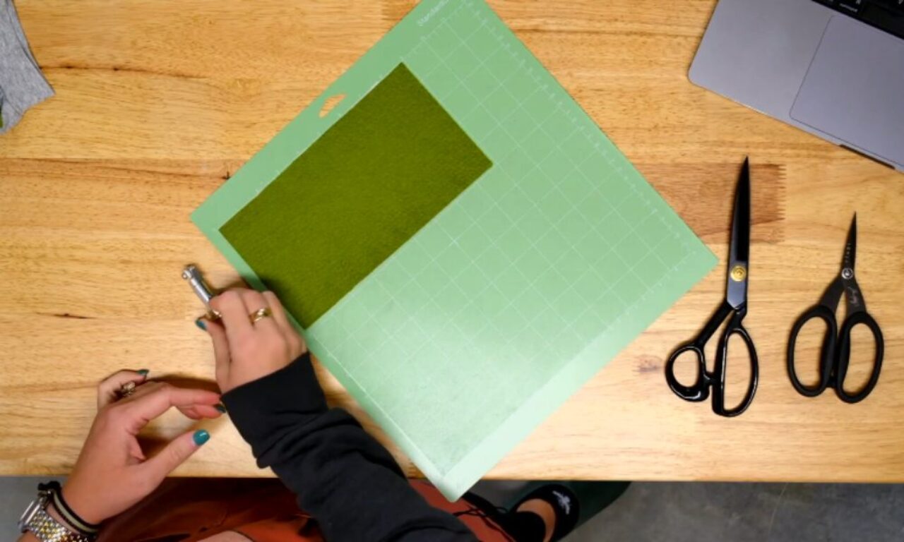 A person is working at a wooden table with a green cutting mat in the center. A rectangular piece of green felt is placed on the mat. The person is holding a precision craft knife in their right hand, preparing to cut the felt. Two pairs of black scissors and the edge of an open laptop are also visible on the table. The person's hands have painted nails and are wearing rings and bracelets.