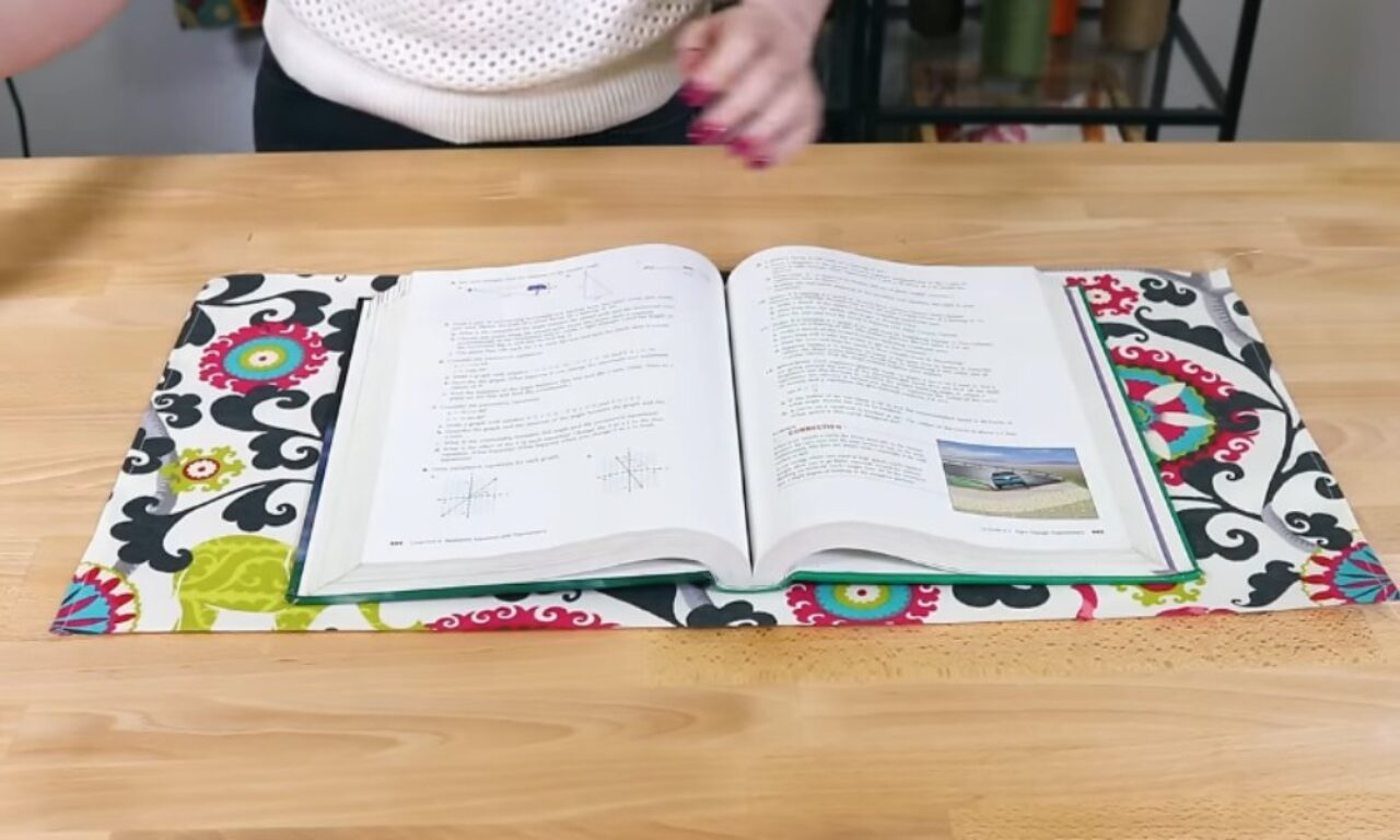 An open textbook or workbook lying on a colorful fabric book cover with a bold floral pattern in black, pink, lime green, and turquoise. The book's pages show some mathematical graphs and text, with a small landscape photo visible on one page. The book is placed on a wooden table surface.