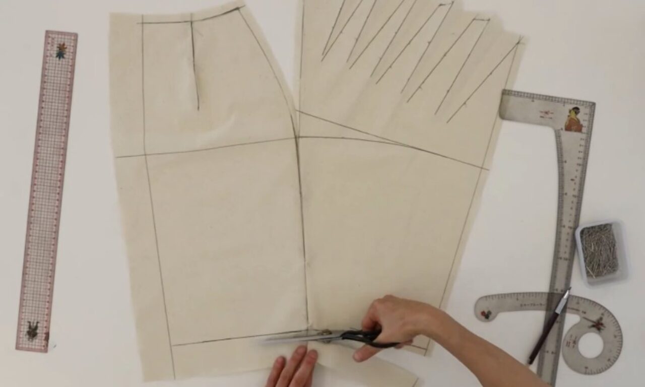 Overhead view of a person's hands using scissors to cut along black lines on a paper sewing pattern for a garment. The pattern is laid out on a white surface, with drafting rulers, a French curve, and pins visible nearby.