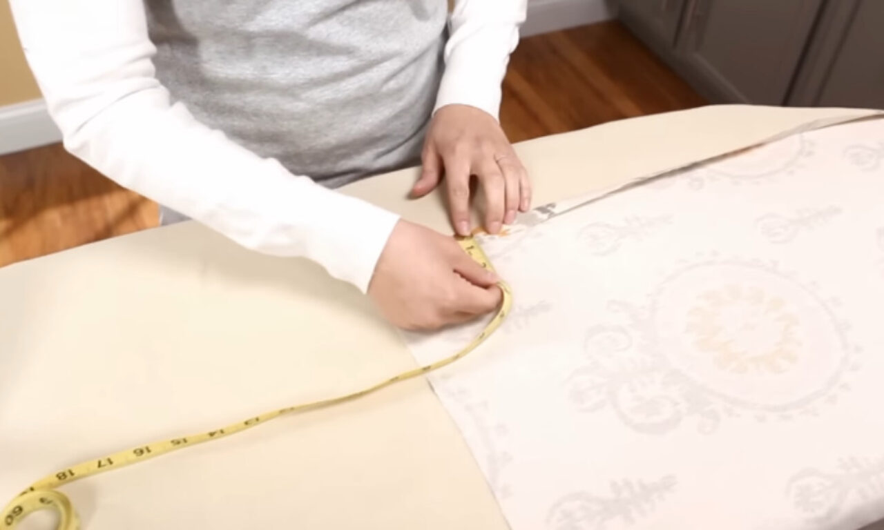 Person measuring fabric with a yellow measuring tape on a table, wearing a gray shirt with white sleeves.