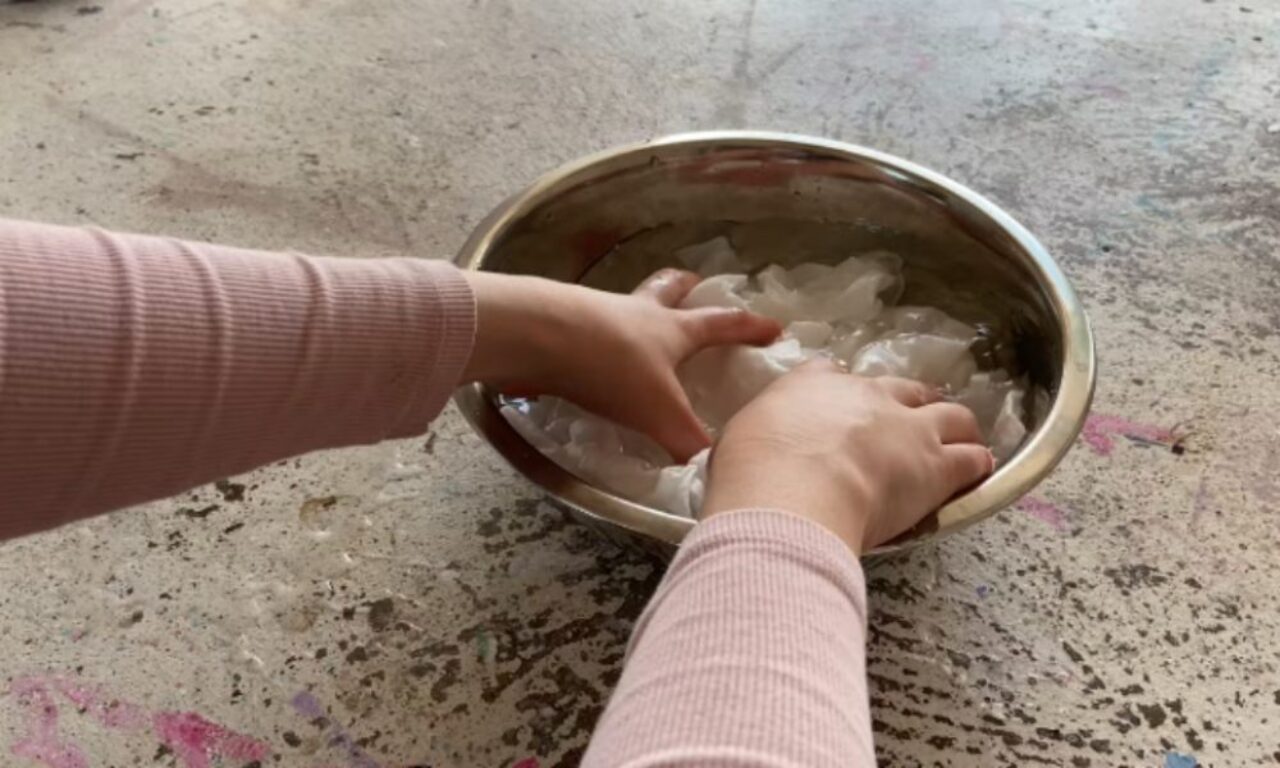 A close-up, slightly overhead view shows a pair of hands in a pale pink ribbed long-sleeved shirt submerging and working with crumpled white material, possibly paper or fabric, in a metal bowl filled with water. The bowl sits on a textured concrete surface with faint paint stains.