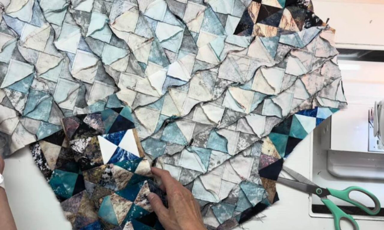 Hands assemble a patchwork quilt of small, irregular blue, white, and grey fabric pieces.