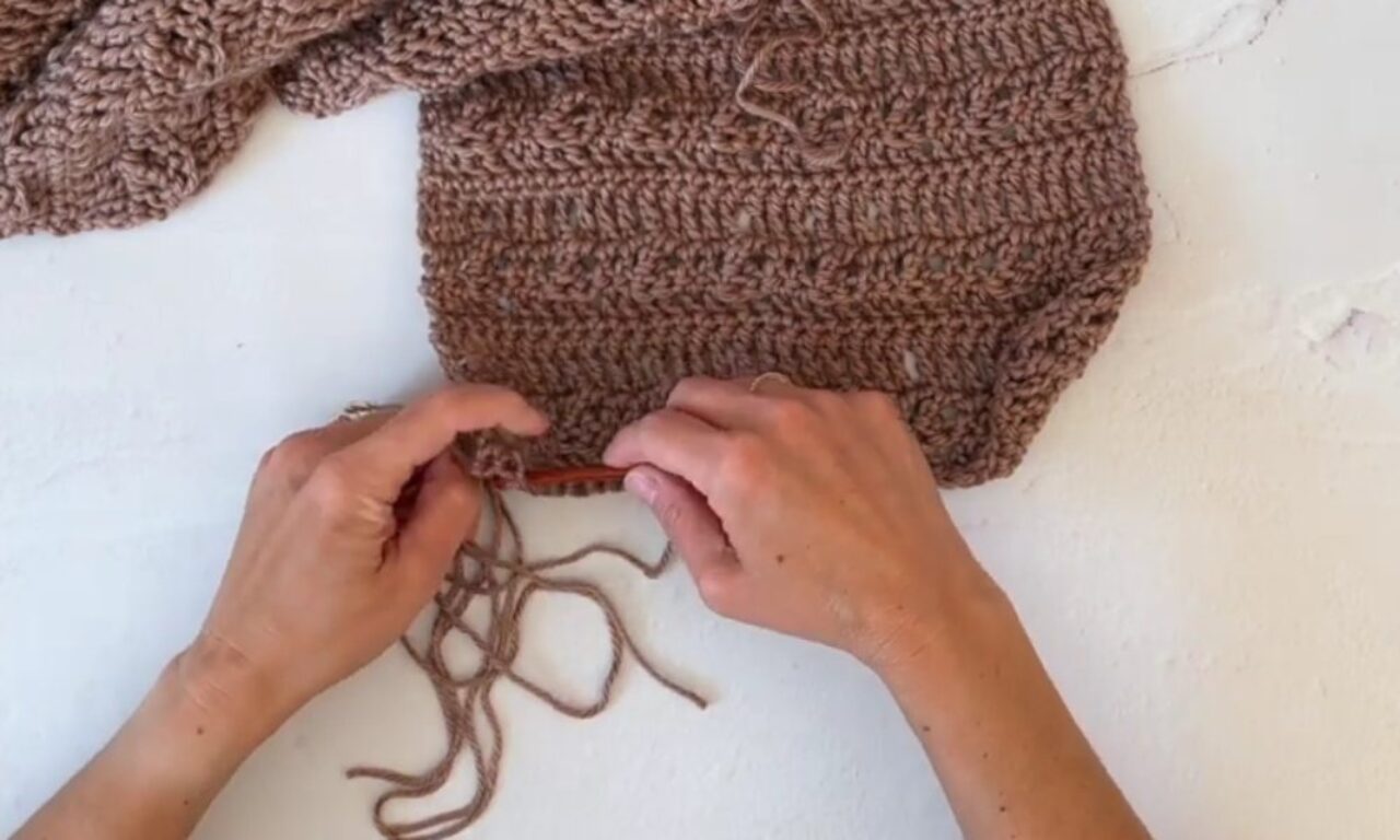 A person's hands crocheting a piece of chunky brown yarn fabric using a crochet hook. The fabric appears to be part of a larger project, possibly a scarf or blanket, resting on a white surface. Loose yarn strands are visible near the working area.