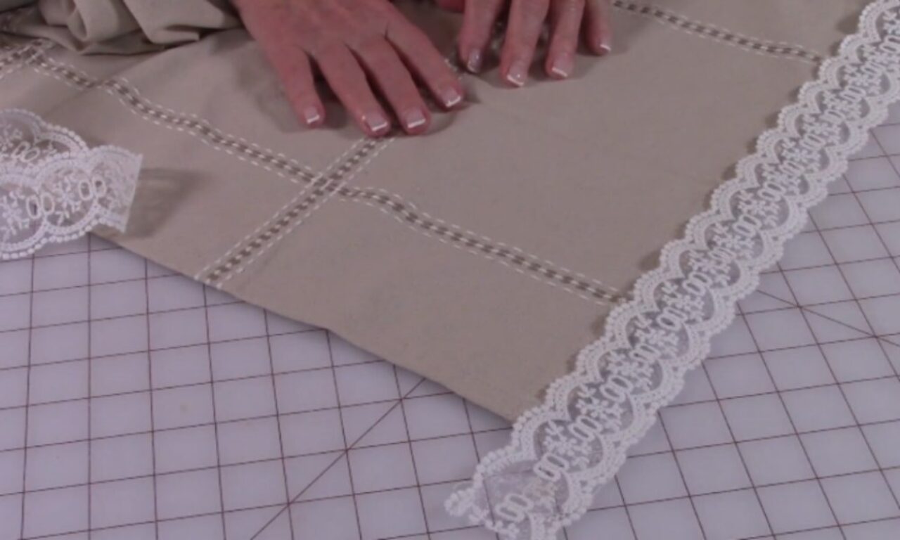 A person's hands are positioned on a piece of beige fabric with decorative stitching forming a grid pattern. A strip of white lace trim is placed along one edge of the fabric, and another piece of lace is visible to the side. The fabric is laid out on a gridded cutting mat, commonly used for sewing or crafting projects.