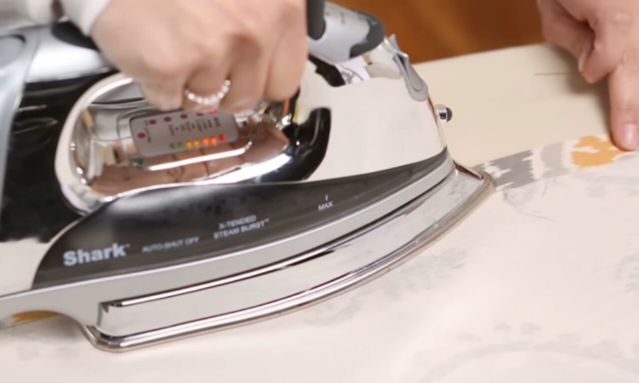 A hand wearing a ring holds a Shark brand iron with the "Auto-Shut Off" feature engaged, pressing it against a piece of fabric with a floral pattern on a light background.