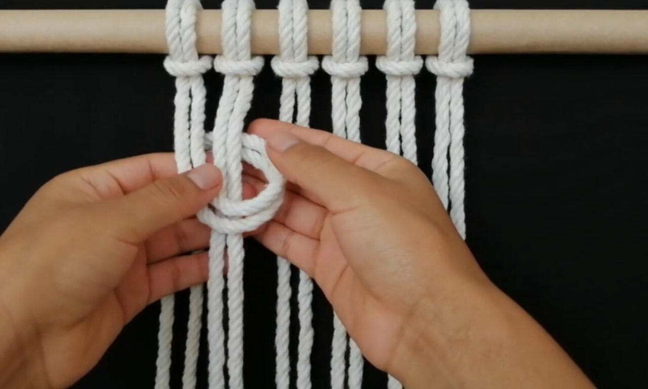 Two hands are shown working with white rope, creating a knot. The rope is hanging from a wooden dowel against a black background. One hand holds a loop of rope, while the other hand guides the strands. The image demonstrates a step in a macramé technique.