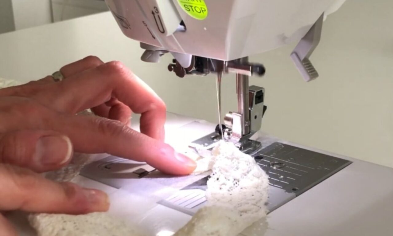Close-up of a person’s hands guiding delicate white lace beneath the moving needle of a sewing machine, illustrating careful, detailed stitching work.