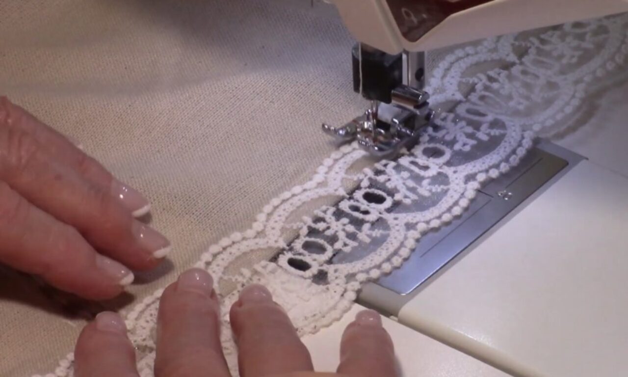 hands guiding a piece of white lace fabric through a sewing machine as it stitches the lace onto a beige textile.