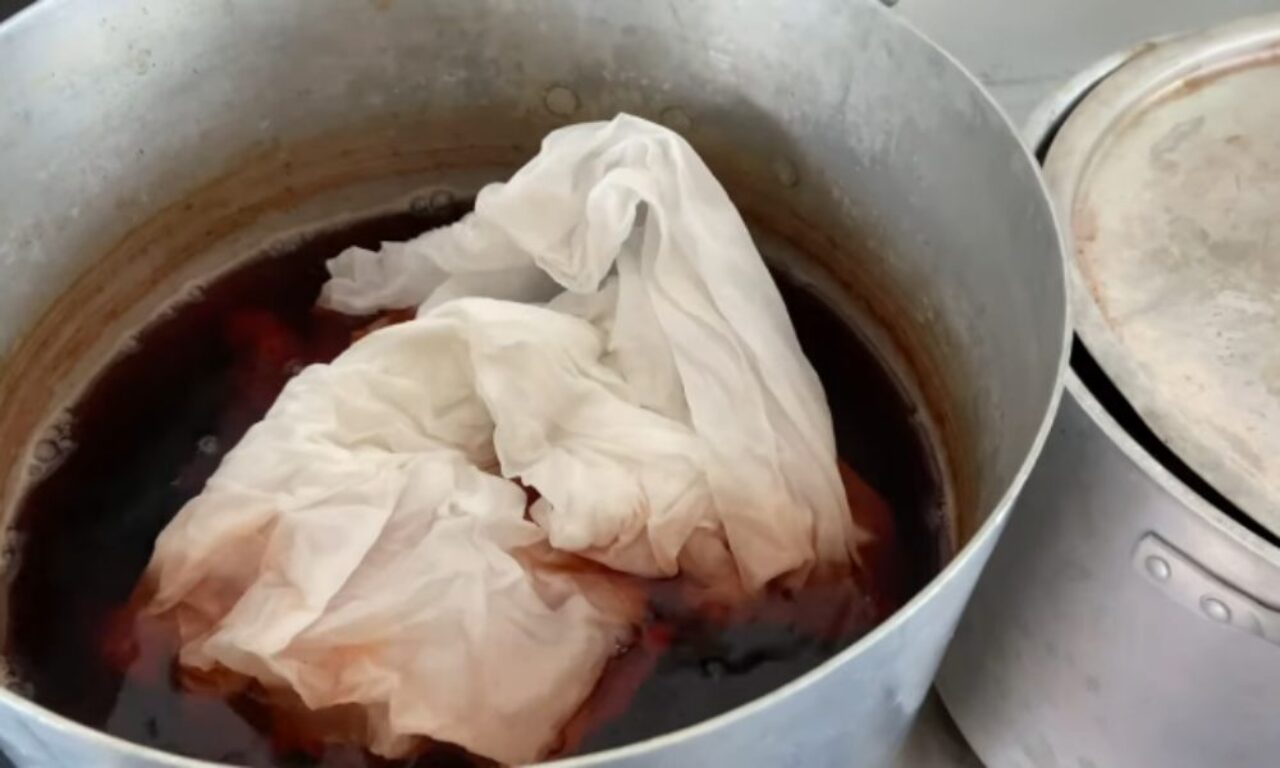 A close-up view into a large metal pot where a white, crumpled piece of fabric is steeping in a dark, reddish-brown liquid, visibly absorbing the color and becoming stained. Another metal pot lid is partially visible to the right.