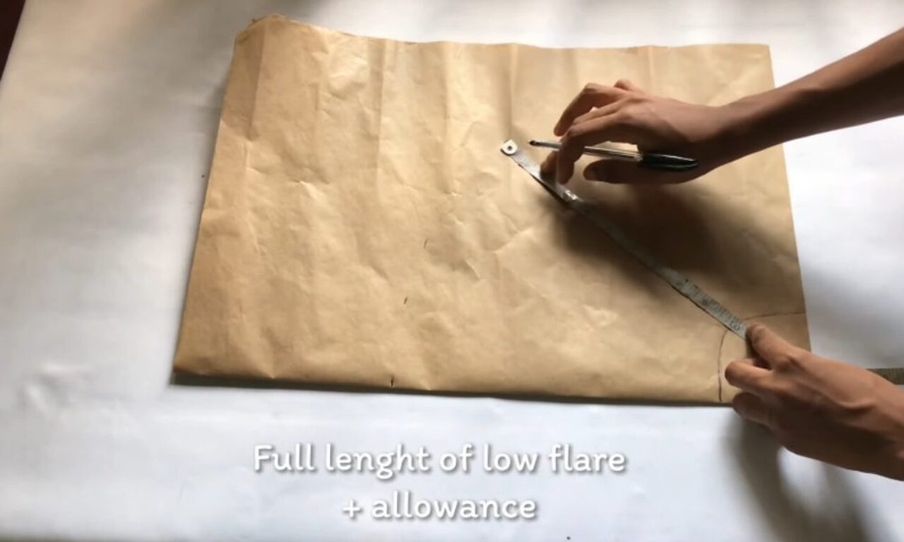 Hands are shown measuring and marking a piece of brown pattern paper with a ruler and pen, preparing for garment making. The text at the bottom of the image reads: "Full length of low flare + allowance."