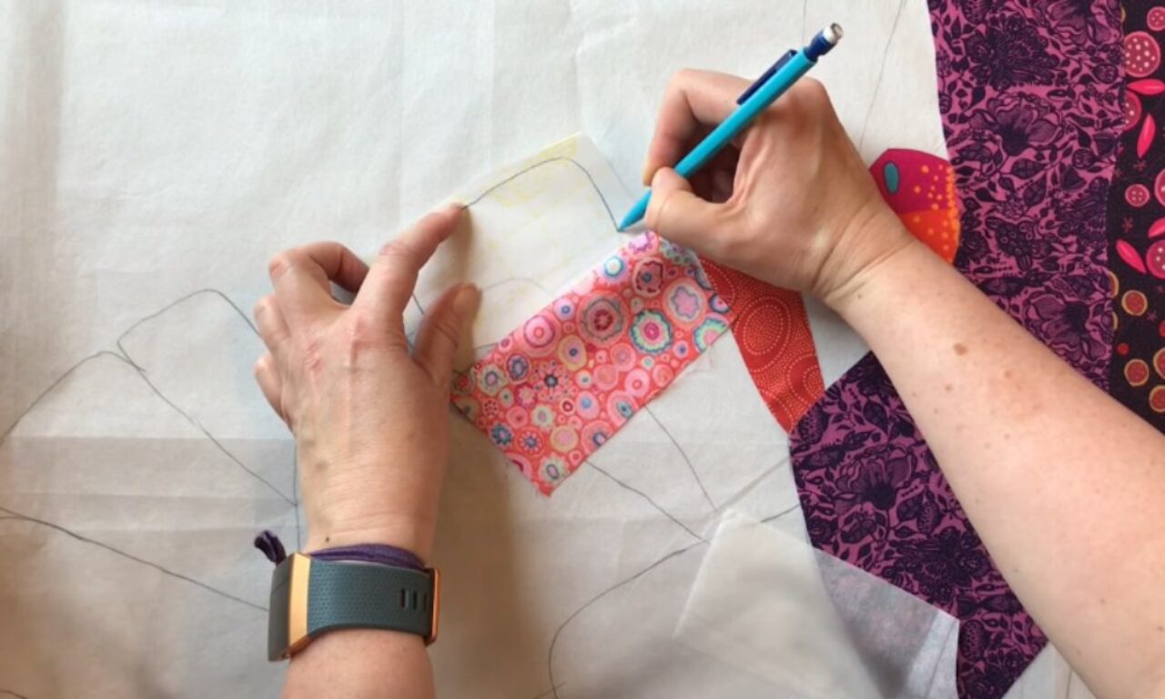A person is working on a quilting project. They are using a blue pen to trace or mark patterns onto a piece of fabric that features colorful, floral designs in shades of pink, red, and purple. The fabric is placed on a white background with faint outlines already drawn on it. The person's left wrist is wearing a fitness tracker or smartwatch. Additional quilt pieces with various patterns can be seen in the background.