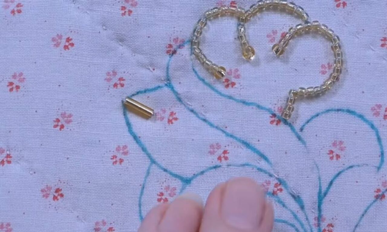 A close-up of a white fabric with small pink flowers and blue embroidery, with a gold chain and two cylindrical gold pieces resting on it. A person's thumb and forefinger are visible at the bottom of the image, with one of the gold pieces between them.