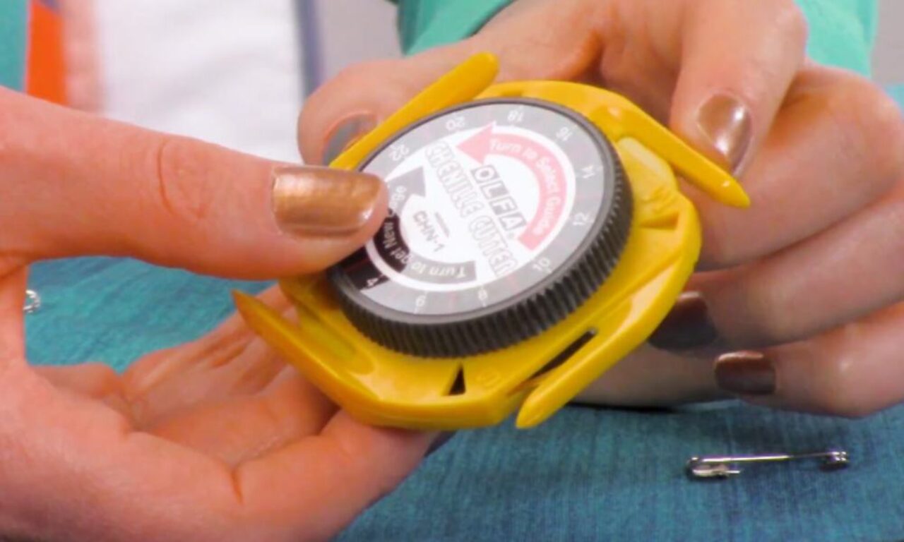A close-up of hands with metallic gold nail polish holding a yellow and black circular object with text and numbers, resembling a small gauge or measuring tool.