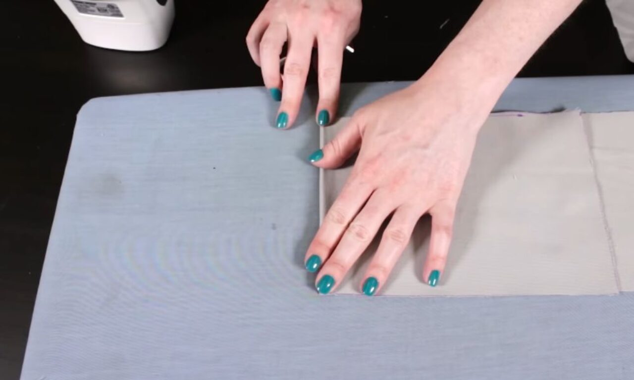 Close-up of two hands with teal nail polish pressing the folded edge of a piece of light grey fabric flat on a blue ironing board, preparing the seam before ironing.