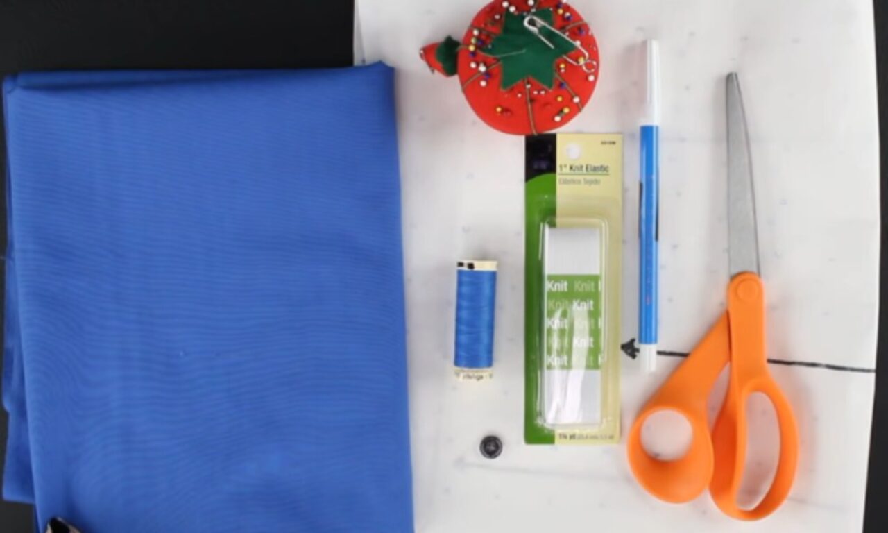 A flat lay of sewing supplies including a folded piece of royal blue fabric, a red tomato-shaped pincushion with multicolored pins, a spool of blue thread, a package of 1-inch knit elastic, a blue fabric marking pen, a single black button, a pair of orange-handled fabric scissors, and a white sewing pattern sheet with faint blue markings.