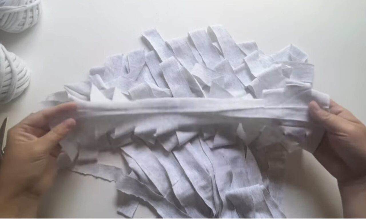 Hands hold a wide white fabric strip horizontally over a pile of short white fabric strips arranged in zigzag layers. Two balls of white fabric yarn are visible in the background.