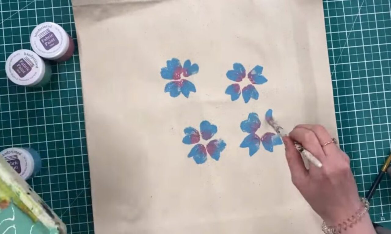 Person painting blue and pink flowers on fabric, with pots of fabric paint visible.